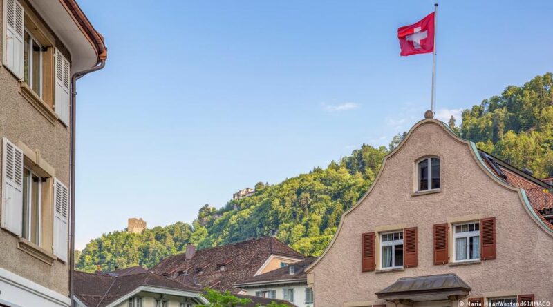 Swiss flag flying atop a beige gabled building, with tiled roofs and green hills in the background