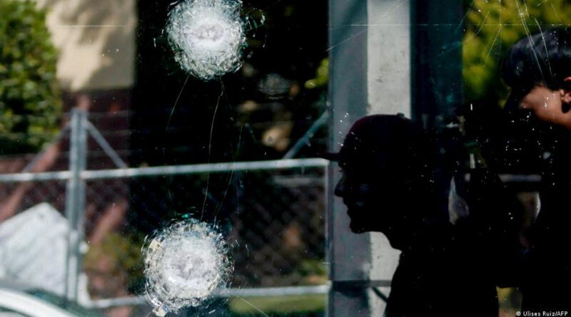 Cracked glass window with two impact holes and dark silhouettes of people reflected outside, suggesting a crime scene welcome to light.