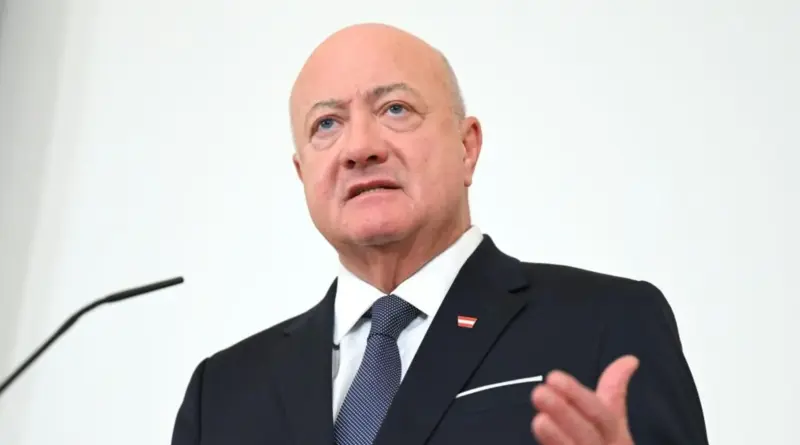 Older man in a dark suit and blue tie speaking at a podium, wearing a small red-and-white flag lapel pin.