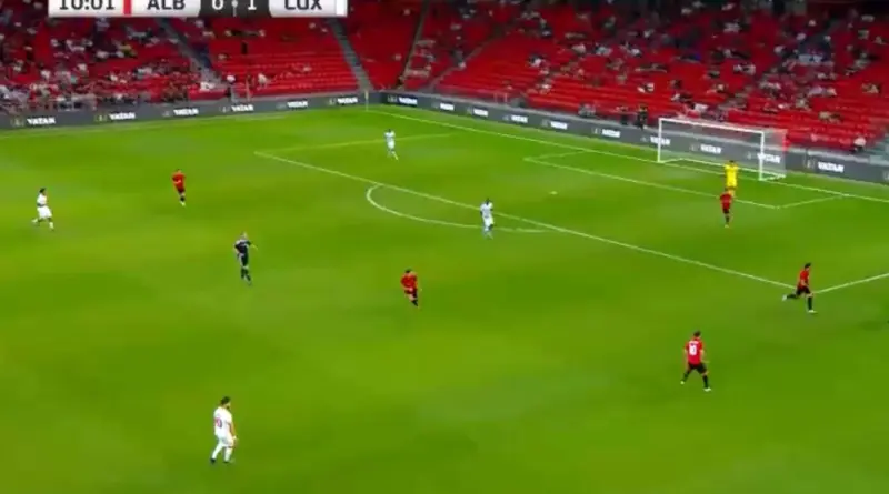 Soccer match in progress on a green pitch; players in white and red kits, goalkeeper in yellow near goal.