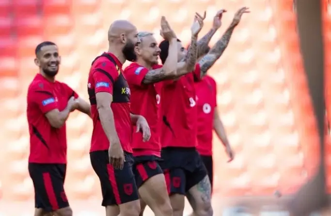 Soccer players in red jerseys celebrate together on the field, some raising their arms in triumph, with orange stadium seats in the background.