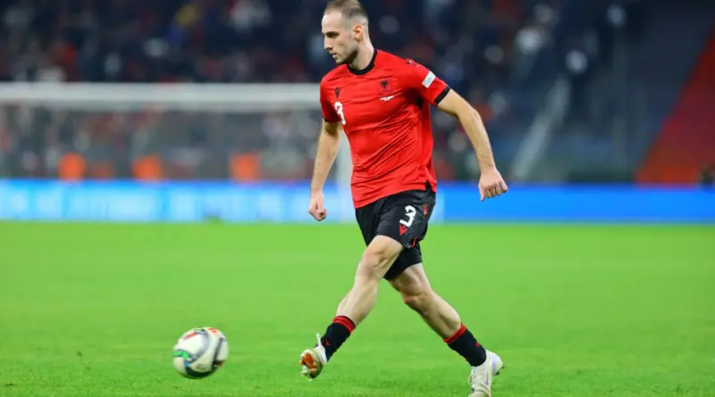 Soccer player in a red jersey and black shorts dribbling a ball on a green field during a match, stadium in background
