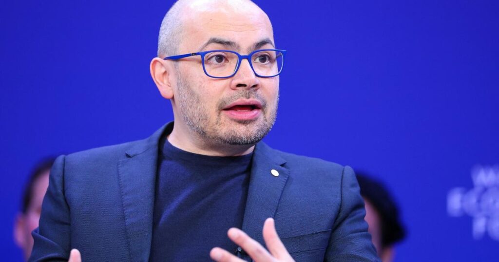 Man wearing blue glasses and a dark blazer speaks during a panel discussion against a blue background, gesturing with his hands.