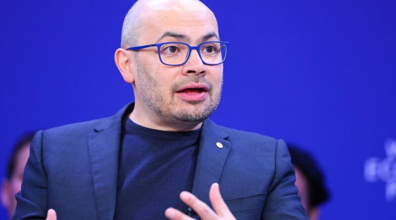 Man wearing blue glasses and a dark blazer speaks during a panel discussion against a blue background, gesturing with his hands.
