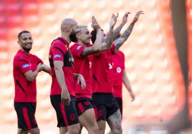 Soccer players in red jerseys celebrate together on the field, raising their arms in a cheer near empty orange seats.
