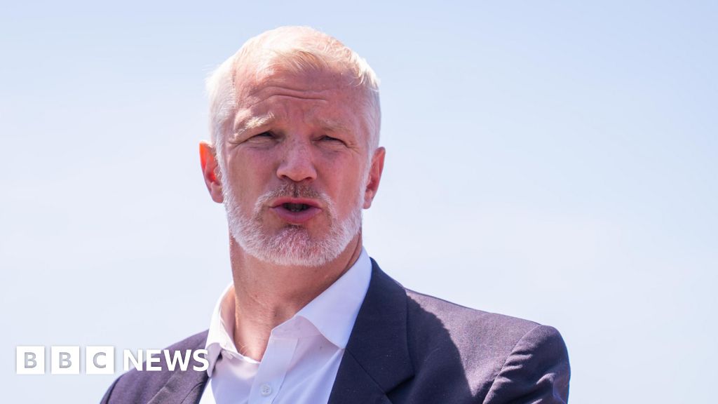 Older man with white hair and beard speaking outdoors in a dark suit and white shirt; clear blue sky behind; BBC News watermark in the lower left corner.