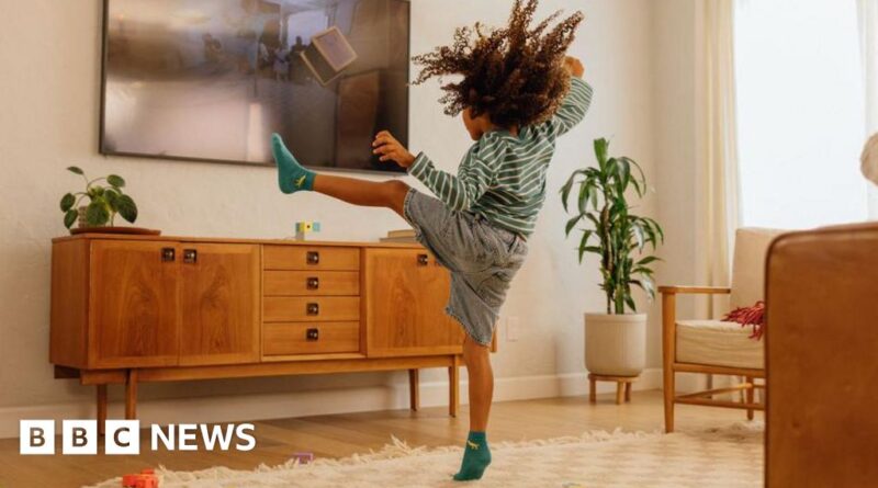 Child in a striped shirt kicks a flat-screen TV mounted on a wall in a cozy living room, wearing teal socks and denim shorts.