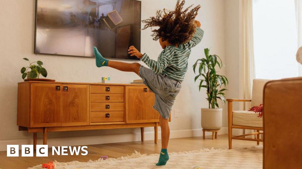Child in a striped shirt kicks a flat-screen TV mounted on a wall in a cozy living room, wearing teal socks and denim shorts.