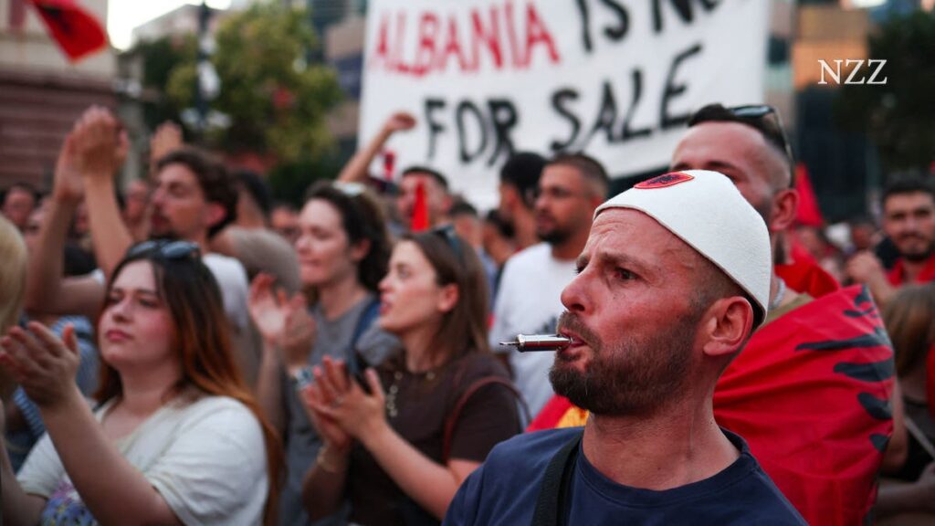 Protest scene with a man in a white cap in the foreground, a pen in his mouth, while a banner behind reads 'Albania is not for sale'.