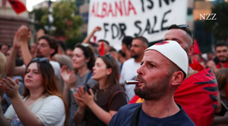Protest scene with a man in a white cap in the foreground, a pen in his mouth, while a banner behind reads 'Albania is not for sale'.