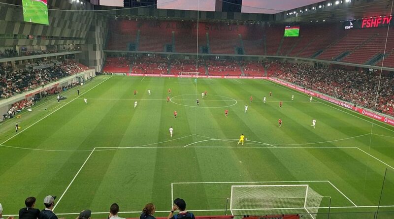 A soccer stadium during a match, view from the stands behind one goal with players on the lush green field and a full crowd in red seats.