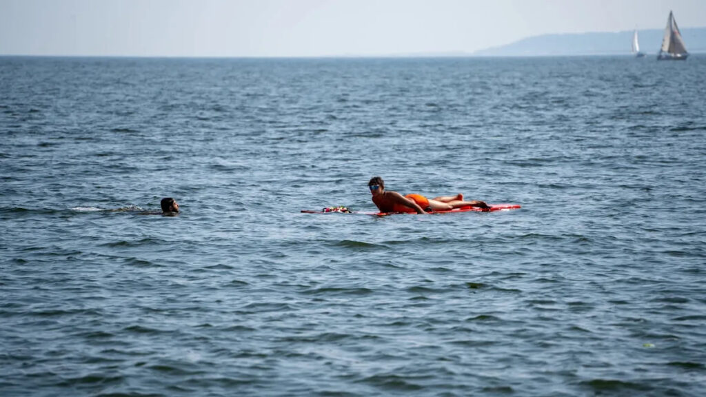 Person in orange swimwear lying on a red bodyboard in the open water, with another swimmer nearby; sailboats on the horizon.