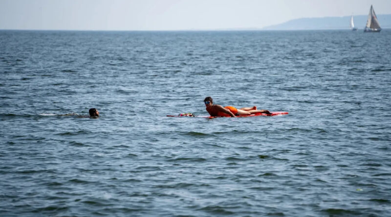 Person in orange swimwear lying on a red bodyboard in the open water, with another swimmer nearby; sailboats on the horizon.