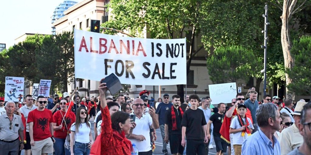 Crowd of protesters marching in a city with a large banner reading 'ALBANIA IS NOT FOR SALE'.