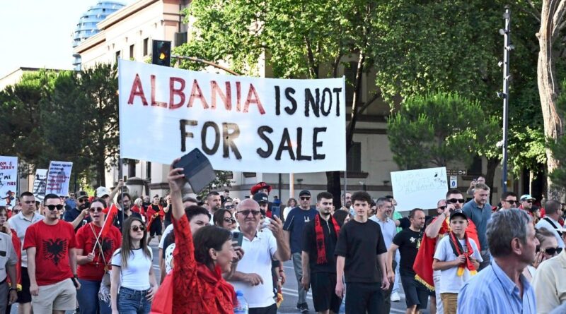 Crowd of protesters marching in a city with a large banner reading 'ALBANIA IS NOT FOR SALE'.