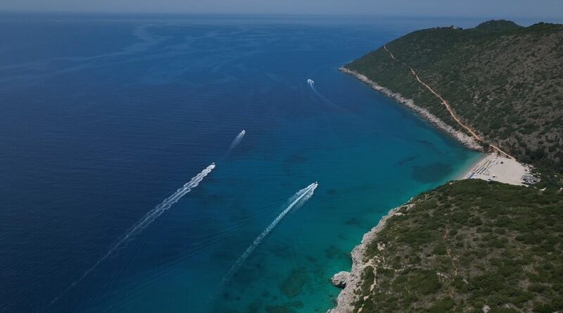 Aerial view of a rocky coastline with turquoise water, a sandy beach on the right, and several speedboats leaving white wakes in the sea.