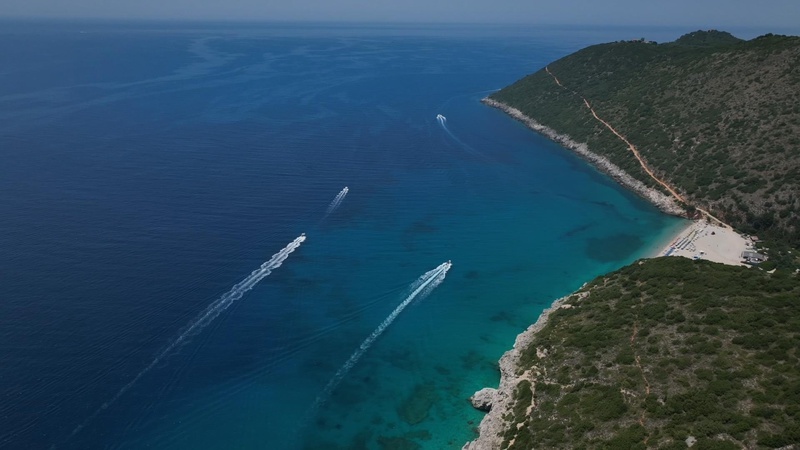 Aerial view of a rocky coastline with turquoise water, a sandy beach on the right, and several speedboats leaving white wakes in the sea.