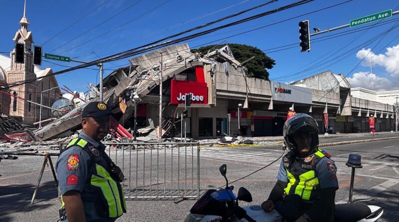 In the foreground, two police officers on motorcycles block off a street with a collapsed building and debris in the background under a clear blue sky.