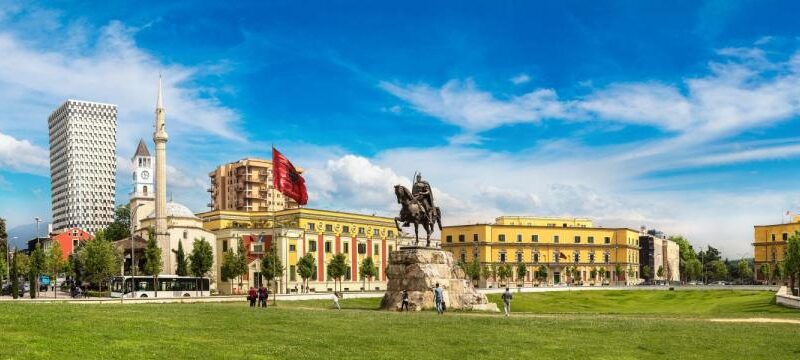 Equestrian statue on a stone pedestal in a city square with yellow buildings and a blue sky behind it.