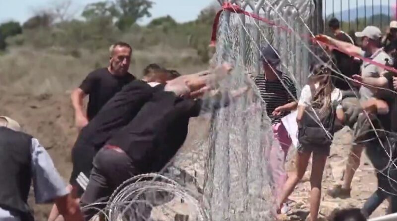 People push against a tall barbed-wire fence in a desert border area, with a line of onlookers on the other side.