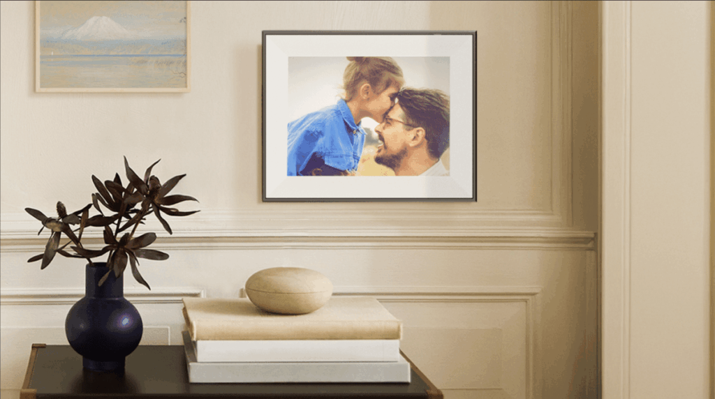 Framed photo of a couple kissing hangs on a beige wall above a side table with a dark blue vase, stacked books, and a smooth stone bowl.