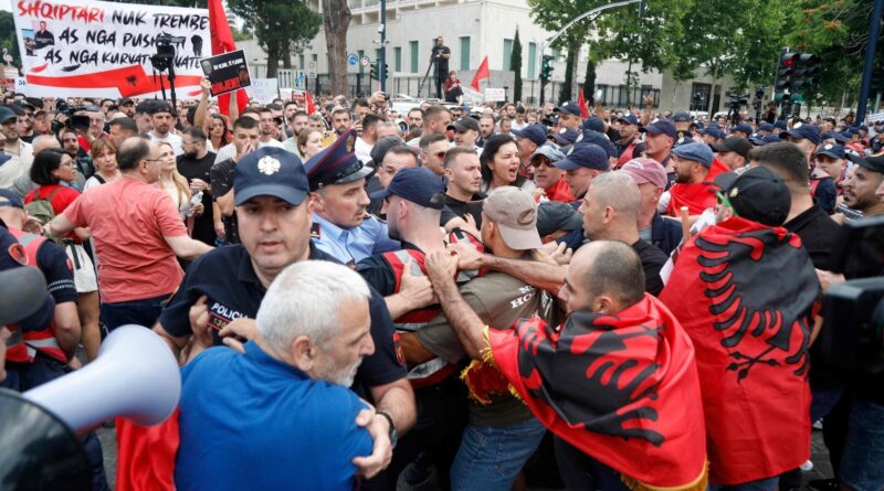 Massive street protest with police in riot gear separating clashing groups; crowd densely packed, many in red jackets and caps, urban background with a large banner.