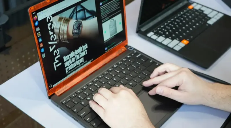 Person typing on an orange‑framed laptop at a white desk, with a second laptop in the background.