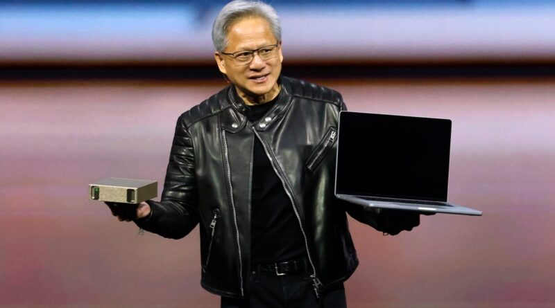 Smiling man in a black leather jacket on stage, holding a small beige device in one hand and a laptop in the other.