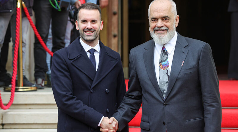 Two men in formal attire shake hands on a red carpet at a ceremony, with onlookers in the background.