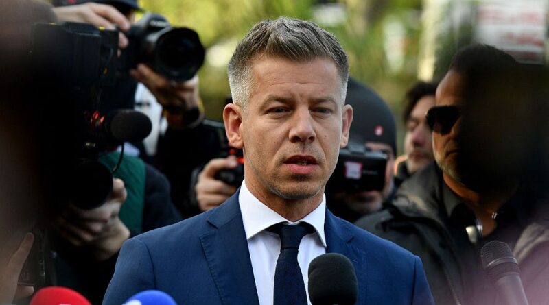 Man in a blue suit speaks to reporters as cameras and microphones surround him outdoors.