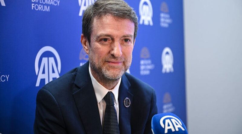 Man in a dark suit speaks into a blue AA microphone at a diplomacy forum press event, with AA logos on the backdrop.