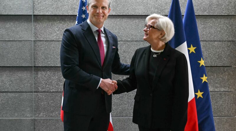 Two adults in business attire shake hands in front of U.S. and European Union flags against a gray stone wall, signaling a formal agreement or meeting.