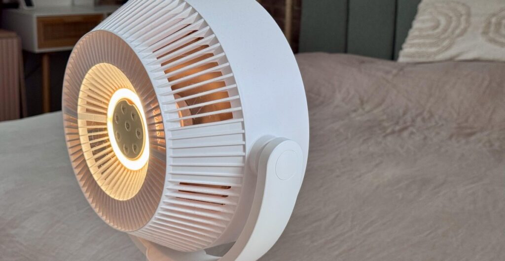White circular heater/fan on a bedspread, glowing warm light at the center, with vertical blade grille design on the housing