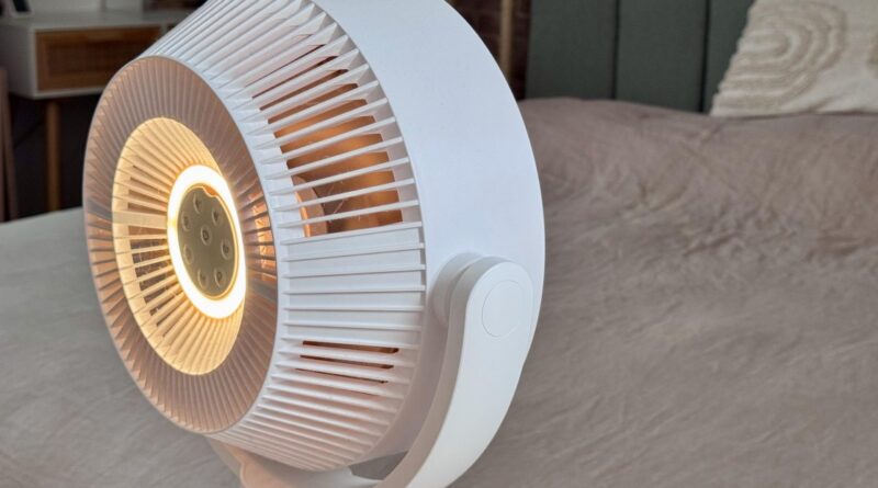 White circular heater/fan on a bedspread, glowing warm light at the center, with vertical blade grille design on the housing