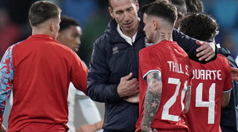 Soccer players in red uniforms surround a coach in a dark jacket, arms around each other in a team huddle outside stadium lights behind them.