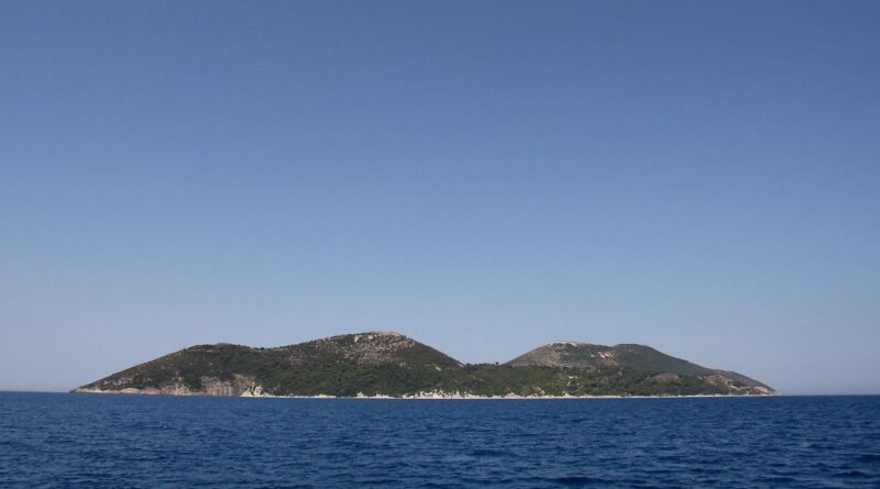 Two small forested islands rising from a calm blue sea under a clear sky, on the horizon in daylight.