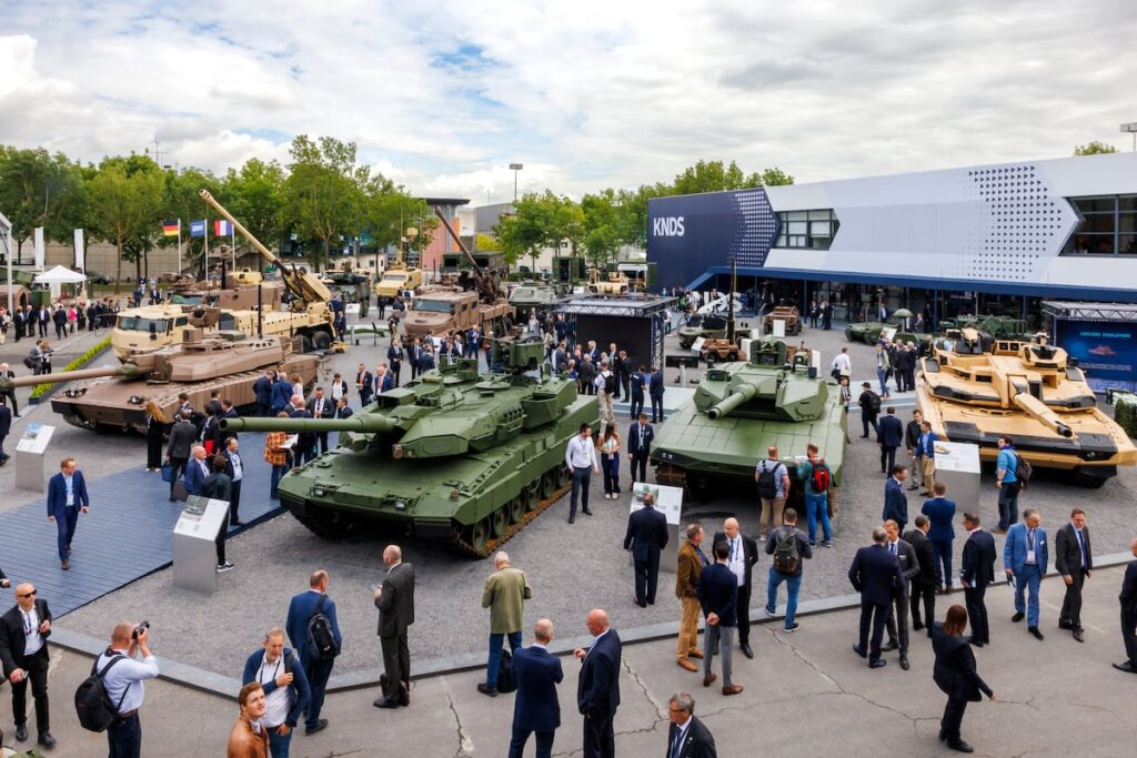 Military exhibition with several tanks and armored vehicles on display, crowds of professionals walking around and conversing near a blue KNDS building.