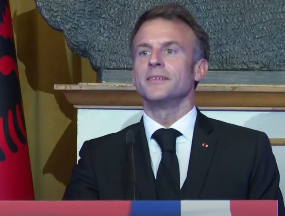 Man in a suit giving a speech at a podium with a blue-red-white banner in front.