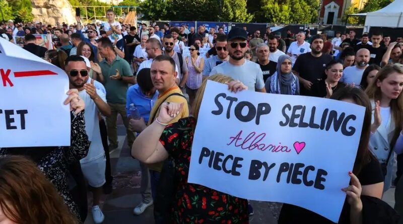 Crowd of protesters at an outdoor rally, holding a sign: 'Stop Selling Albania Piece by Piece' with a heart symbol in the foreground.