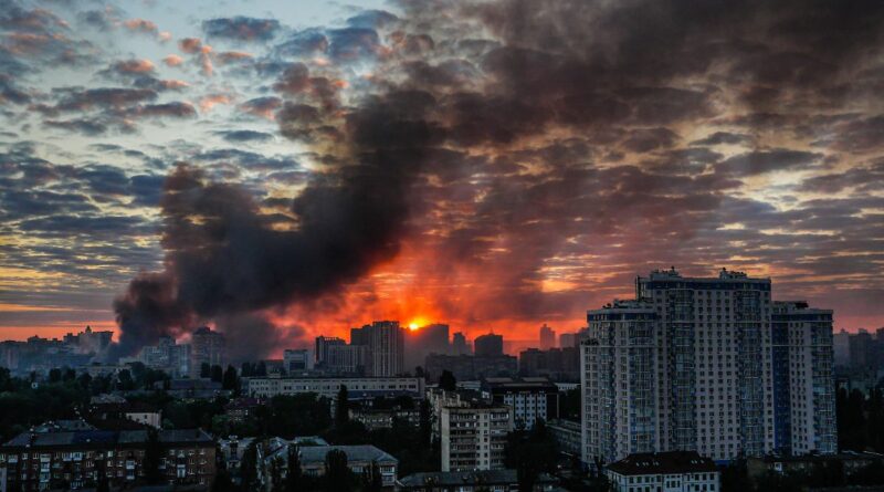 City skyline at sunset with a large plume of black smoke rising from a fire near the horizon.