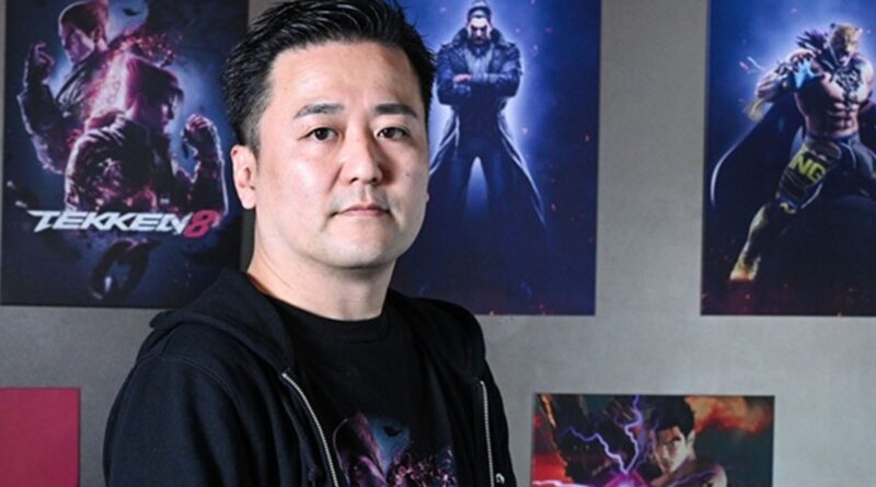 Portrait of a man in a dark hoodie standing in front of Tekken 8 posters on a wall.