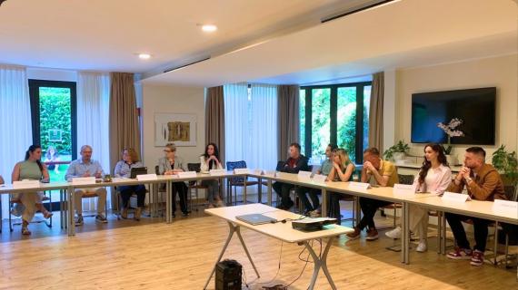 A group of people seated around a U‑shaped conference table in a bright room, discussing with a projector on a small stand in front.