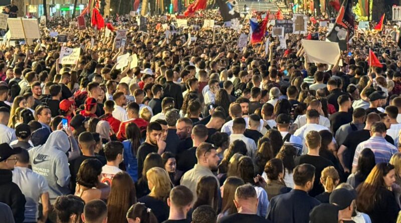 Crowd of protesters filling a city street at dusk, holding signs and flags amid streetlights.