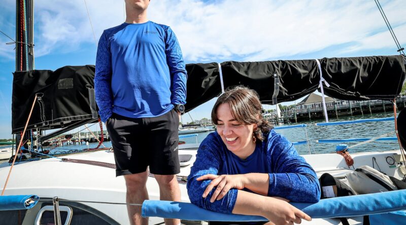 Two people in blue shirts on a sailboat; man stands with hands in pockets while woman sits at the edge smiling by the railing.
