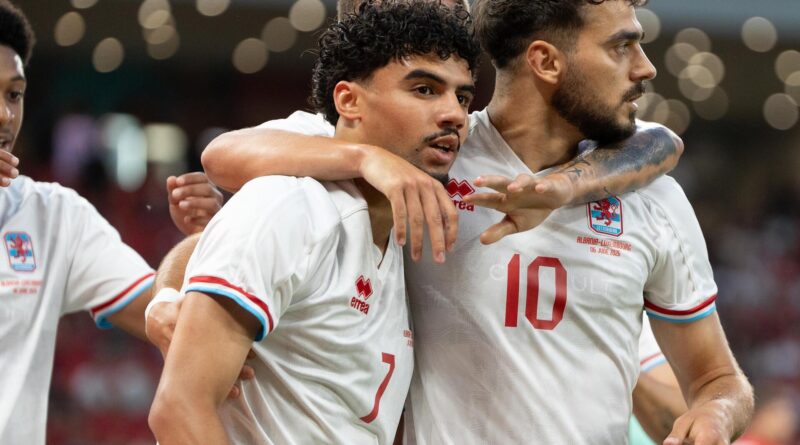 Three soccer players in white jerseys celebrate, arms around each other in a group hug, smiling faces visible in a stadium setting.