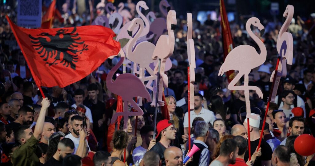 Crowd at a rally holding large pink flamingo cutouts on sticks and a red Albanian flag with a black eagle.