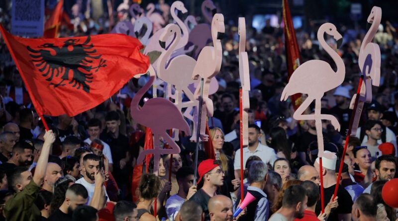 Crowd at a rally holding large pink flamingo cutouts on sticks and a red Albanian flag with a black eagle.