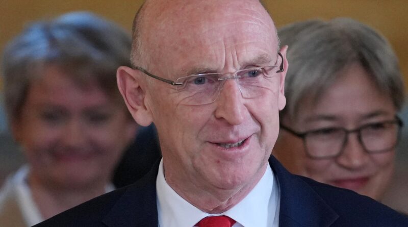 Close-up of an older man with rimless glasses, wearing a dark suit and red tie at a formal event, smiling slightly.