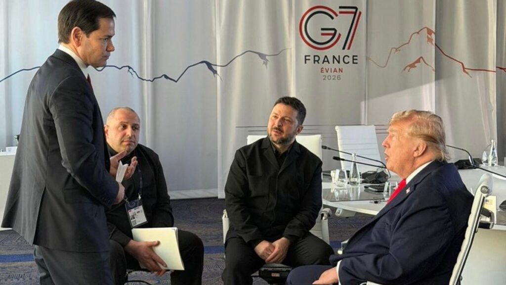 Four men in a meeting room: one standing in a suit talks to three seated colleagues, with a France Evian 2026 banner in the background.