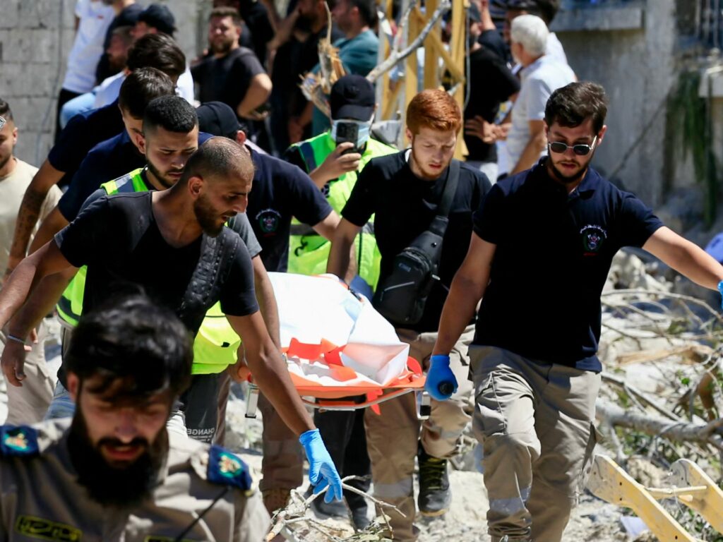 Rescue workers in uniforms and gloves carry a person on a stretcher through rubble at a disaster site.